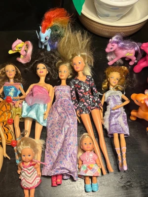 Barbiedockor & My Little Pony-leksaker - Samling med flera Barbiedockor i olika klänningar och två mindre dockor samt fem My Little Pony-figurer i olika färger. Dockorna har kläder i lila, rosa, blått och mönstrat tyg. Ponysarna är lila, rosa, blå, orange och vit med färgglada manar.