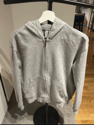 Grå zip-hoodie från Divided  - Klassisk grå zip-hoodie från Divided med dragkedja framtill, två stora fickor och huva med dragsko. Tillverkad i mjukt sweatshirtmaterial, perfekt för en avslappnad stil. Ribbade muddar vid ärmslut och nederkant.
