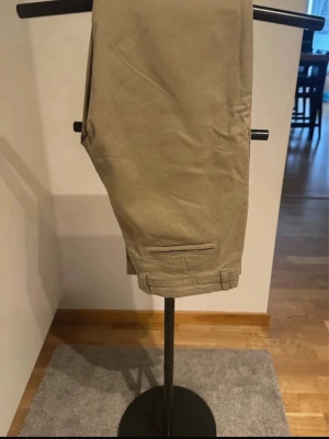 Beiga Dobber chinos 32 - Dobber chinos för herr i storlek 32. Byxorna är beige och tillverkade i bomull och elestan. Klassisk modell med hällor för bälte, sidofickor fram och passpoalerade bakfickor.