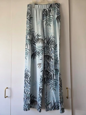 Ljusblå vida loungewear-byxor med blad - Superbekväma loungewear-byxor i ljusblått med svart och grått bladmotiv. Byxorna har resår i midjan och är vida i modellen, perfekta för chill dagar hemma. Materialet känns mjukt och svalt mot huden.