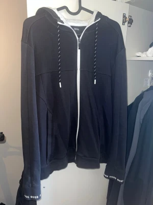 Hugo Boss Hoodie - Snygg svart hoodie från Hugo Boss med dragkedja framtill och vita detaljer på snörena. Broderad logga på bröstet och BOSS-tryck på ärmsluten. Mjuk bomullskänsla och klassisk passform med huva.