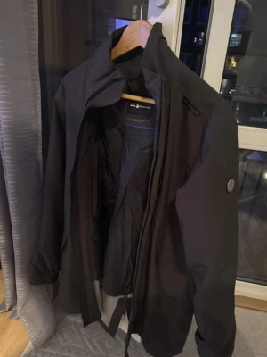 Svart vindjacka Sail racing  - Snygg svart vindjacka från Sail Racing med hög krage och dragkedja. Jackan har en modern passform, innerficka med dragkedja och diskret logga på ärmen. Tillverkad i ett lätt och slitstarkt material som står emot vind.