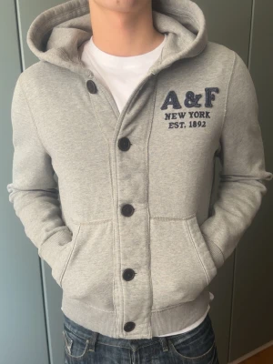 Grå hoodie med knappar från A&F - Säljer en Abercrombie & Fitch hoodie i fint skick. Inga defekter, knappt använd och fortfarande väldigt fräsch. Perfekt som en stilren och bekväm basic till vardags.  Modellen på bilden är 187 cm lång och väger 80 kg för referens på passform.  Snabb leverans 🚚MÅTT: axelmått: 49 cm längd: 64 cm byst: 57 arm(under) 56 arm(över) 71 