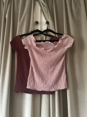Två smockade toppar i rosa och vinrött - Två kortärmade toppar med smockad struktur, en i ljusrosa och en i vinröd. Båda har en söt, vågig kant vid halsringningen och är tillverkade i ett stretchigt material. Perfekta att styla med jeans eller kjol. Den vi röda har lappen kvar medan den rosa är använd 1 gång.