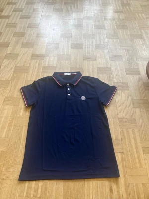 Marinblå Moncler pikétröja - Snygg marinblå pikétröja från Moncler med klassisk krage och tre knappar framtill. Knappt använd och växt ifrån den.