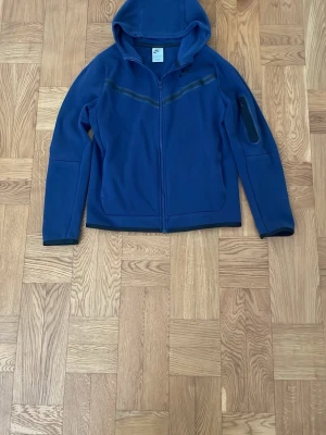 Blå hoodie med dragkedja från Nike - Snygg blå hoodie från Nike med hel dragkedja och huva. Har svarta detaljer över bröstet och på ärmen samt en diskret Nike-logga på bröstet. Bra skick utöver litet märke på baksidan av ena ärmen.