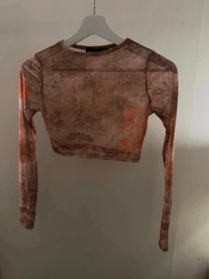 Brun mesh croppad långärmad topp PrettyLittleThing - Trendig croppad mesh-topp i bruna och orange toner med diskret mönster. Långärmad och tight passform, 