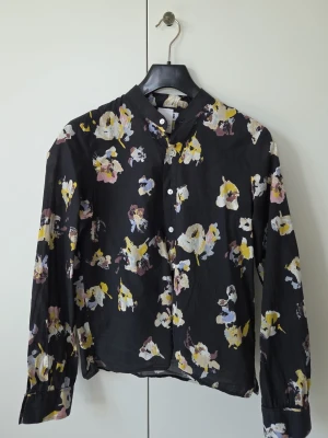 Svart blommig skjorta med knappar - Svart skjorta med blommigt mönster i gult, vitt, rosa och lila. Skjortan har klassisk krage, knäppning framtill och långa ärmar. Materialet är tunt och luftigt, perfekt för en trendig look.