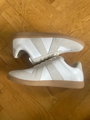 Maison Margiela gats skor - Stilrena maison margiela gats sneakers i vitt och beige med ovandel i slätt läder och detaljer i mocka. Skorna har klassisk gummisula i ljusbrunt och matchande vita skosnören. Perfekta för en clean och modern look.