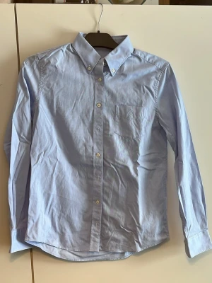 Ljusblå skjorta med knappkrage - Klassisk ljusblå skjorta med button-down krage och bröstficka. Skjortan har långa ärmar och vita knappar framtill. Perfekt till både jeans och chinos för en clean look.