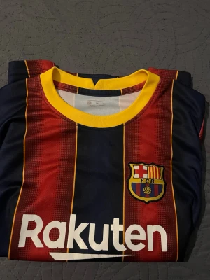 FC Barcelona Ronaldinho matchtröja - Säljer en klassisk FC Barcelona matchtröja med Ronaldinho och nummer 10 på ryggen. Tröjan har rödblå vertikala ränder, gula detaljer och klubbmärke på bröstet. Tillverkad i lätt och ventilerande material, perfekt för fotboll eller som supporterplagg.