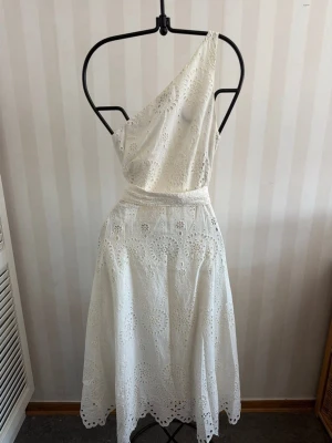Vit oneshoulder klänning med broderi - Säljer en vit oneshoulder klänning med vackra broderade detaljer och hålmönster. Klänningen har en markerad midja och ett svepande fall som ger en elegant siluett. Perfekt för sommarens alla tillfällen!
