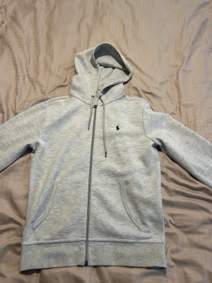 Två hoodies från Polo Ralph Lauren - Säljer två snygga hoodies från Polo Ralph Lauren, en i mörkblått och en i ljusgrått. Båda har dragkedja, huva med snören och klassisk Polo-logga på bröstet. Perfekt för chill dagar och streetstyle. Materialet är mjukt och bekvämt, med stora fickor framtill.