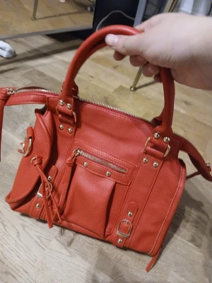 Röd handväska med gulddetaljer - Cool röd handväska i skinnimitation med guldfärgade metalldetaljer, dragkedja upptill och flera fickor framtill. Väskan har två rejäla handtag och dekorativa spännen. Perfekt för dig som vill sticka ut med en färgstark accessoar.