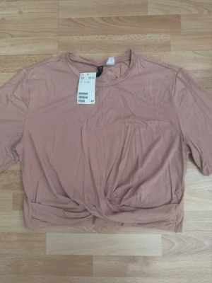 Ny rosa croppad topp från H&M - Trendig  croppad topp från H&M med twistad detalj framtill. T-shirten har korta ärmar och rund halsringning, tillverkad i mjuk bomull. Perfekt för en avslappnad och stilren look.