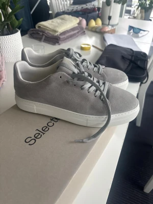 Gråa sneakers  - Snygga gråa sneakers från Selected med ovandel i mjuk mocka och vita, tjocka sulor. Klassisk låg modell med matchande grå skosnören och diskret design. Perfekta för dig som gillar stilrena och minimalistiska skor. Endast testade en gång