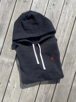 Svart hoodie från Polo Ralph Lauren - Svart hoodie från Polo Ralph Lauren med vit dragsko och klassisk röd broderad logga på bröstet. Mjuk bomullskänsla och stor huva. Perfekt för en avslappnad och stilren look.