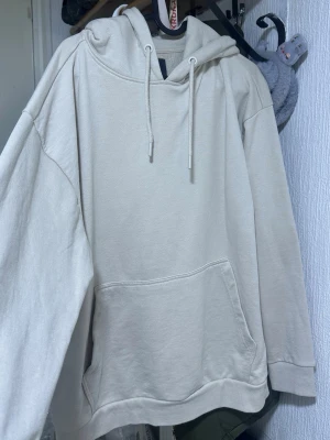 Beige oversized hoodie med ficka - En stilren beige hoodie med huva och dragsnören. Modellen är oversized med en stor magficka framtill och ribbade muddar vid ärmslut och nederkant. Perfekt för en avslappnad och trendig look.