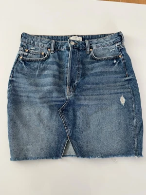 Blå jeanskjol med slits och rå kant - Snygg blå jeanskjol i klassisk femficksmodell med slits framtill och rå, fransig nederkant. Kjolen har en lätt tvättad look och slitningar för en trendig vibe. Perfekt till sneakers eller boots för en avslappnad stil.
