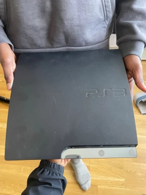 PlayStation 3 Slim svart konsol - Säljer en svart PlayStation 3 Slim från Sony. Konsolen har en stilren, matt finish och är tillverkad i plast. Den klassiska PS3-loggan syns på ovansidan och designen är rektangulär med rundade kanter. Perfekt för dig som vill spela klassiska PS3-spel.