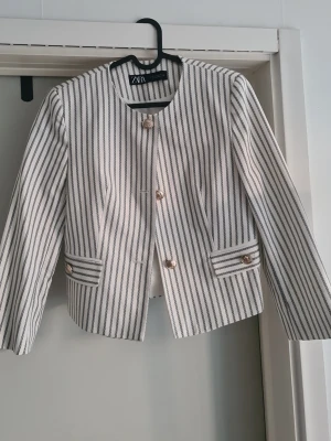 Randig kavaj från Zara med guldknappar - Snygg kavaj från Zara i vitt med svarta smala ränder och coola guldknappar framtill. Rak, kortare modell med rund halsringning och dekorativa fickdetaljer. Perfekt för en trendig och stilren look.