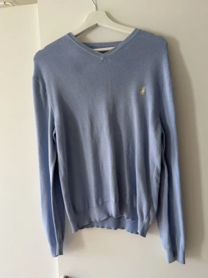 Ljusblå tröja från Ralph Lauren - Ljusblå långärmad tröja från Ralph Lauren med v-ringning och diskret broderad logga på bröstet. Tröjan är tunnstickad och har ribbade muddar vid ärmslut och nederkant. Perfekt för en stilren och avslappnad look.