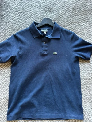 Marinblå pikétröja från Lacoste - Klassisk marinblå pikétröja från Lacoste med korta ärmar och krage. Tröjan har knappar framtill och den ikoniska gröna krokodilloggan på bröstet. Perfekt för en stilren och sportig look.