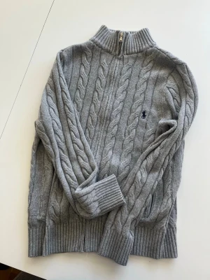  zip-tröja Polo Ralph Lauren - Snygg grå kabelstickad tröja från Polo Ralph Lauren med zip framtill och ribbade muddar. Tröjan har ett diskret broderat logotyp på bröstet och är tillverkad i mjukt material. Storlek M men passar S också