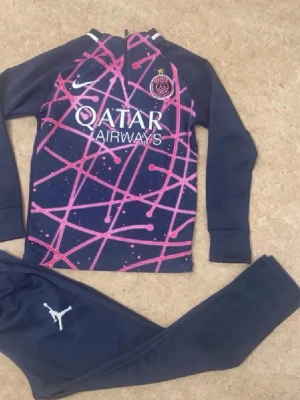 PSG träningsset navy/pink Nike Jordan - Snyggt PSG träningsset i navy med rosa och lila grafiskt mönster på tröjan. Setet har vita loggor från Nike och Jordan samt Qatar Airways tryck på bröstet. Tröjan har lång ärm och dragkedja vid halsen, byxorna är smala och har Jordan-logga. Perfekt för fotbollsträning.