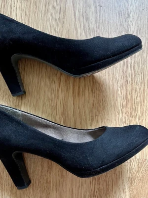 Svarta pumps från Tamaris - Stilrena svarta pumps från Tamaris med rundad tå och klack i klassisk modell. Mockaimitation med  en mjuk innersula. Ca  7 cm klackhöjd. Smala i passformen. Använd endast två gånger. 