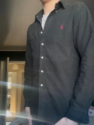 Svart skjorta från Ralph Lauren - Klassisk svart skjorta från Ralph Lauren med vit knappstängning och den ikoniska röda loggan broderad på bröstet. Skjortan har lång ärm och normal passform, perfekt till jeans eller chinos. Tillverkad i mjuk bomull för en skön känsla.