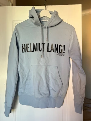 Ljusblå Helmut Lang hoodie - Ljusblå hoodie från Helmut Lang med stor svart logga på bröstet. Sparsamt använd, har däremot en missfärgning i luvan (kolla bild) som kom efter första tvätten. Inget som man tänker på när man har den på sig dock. 