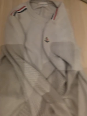 Beige stickad tröja från Moncler - Snygg beige stickad tröja från Moncler med rund halsringning och långa ärmar. Tröjan har Moncler-logga på ärmen och coola ränder i rött, vitt och blått på axlarna. Perfekt för dig som gillar stilrena och sportiga detaljer.