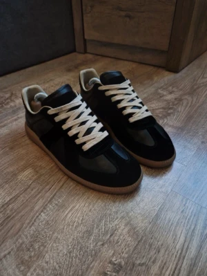 Maison margiela gats - Snygga svarta sneakers med beige sula och vita snören. Ovandelen är i en mix av mocka och läder, vilket ger en stilren och klassisk look. Insidan är ljusbeige och skon har en låg profil som passar till många outfits.