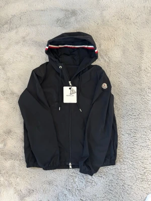 Svart vindjacka från Moncler - Snygg svart vindjacka från Moncler med huva och dragkedja framtill. Jackan har vita och röda detaljer på huvan samt Moncler-logga på ärmen. Justerbar dragsko i huvan och elastiska muddar vid ärmslut och midja. Perfekt för blåsiga dagar.