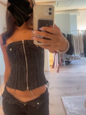 Svart jeans korsett med dragkedja - Säljer en svart korsettopp i denim med dragkedja framtill. Toppen är axelbandslös och har  knappar upptill. Perfekt för dig som vill ha en edgy och trendig look.