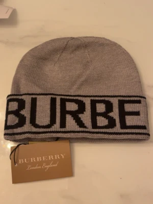 Grå mössa från Burberry med logga - Snygg grå stickad mössa från Burberry med stor svart logotyp runt kanten. Klassisk modell i mjukt material som håller dig varm. Perfekt accessoar för att lyfta din outfit med en exklusiv touch.