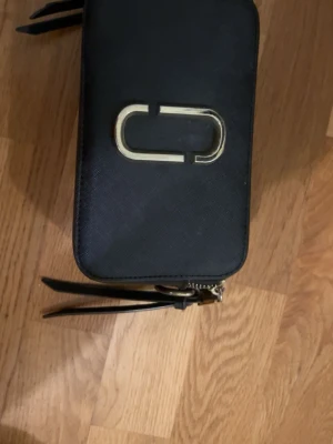Svart Marc Jacobs Snapshot väska - Snygg svart handväska från Marc Jacobs med rektangulär form och dubbla dragkedjor i guld. Väskan har en tygbit som separerar det två komparitmenten emellan men som har lossnat på toppen och istället inte separerar båda delarna men det syns inte utifrån. Väskan har även lite smink fläckar inne i den men det är inget som syns på det yttre. Väskan rymmer mycket plats och jätte snygg till sommaren.
