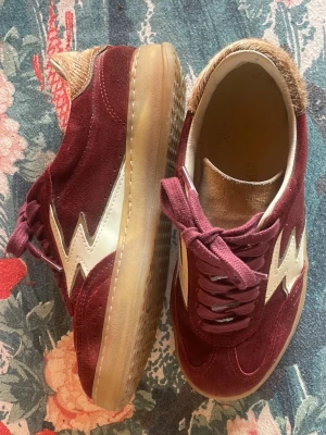 Moa Concept sneakers - Snygga vinröda sneakers från Good News med vit blixtdetalj på sidan. Ovandelen är i mocka och skon har en genomskinlig gummisula. Hälpartiet har en beige textilpanel som kontrast. Perfekta för dig som vill sticka ut med din stil. Helt nya, använda ett fåtal gånger