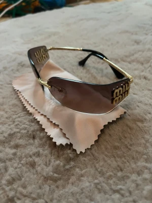 Miu Miu solglasögon med guldiga detaljer - Snygga solglasögon från Miu Miu med bruna tonade glas och guldfärgade metallbågar. Inga synliga repor. Säljer billigt då jag tappat bort fodralet så att man endast får med putsduk och har ej kvar kvitto!