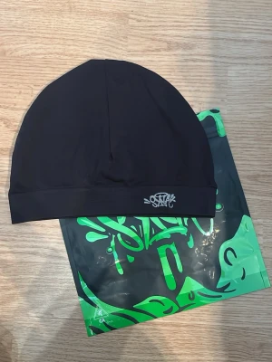 Svart Syna World mössa - Snygg svart mössa från SYNA WORLD med en liten broderad logga framtill. Tillverkad i mjukt och stretchigt material som sitter skönt på huvudet. Inte använd.