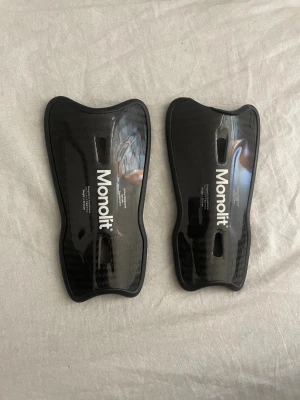Svarta benskydd från Monolit - Säljer ett par svarta benskydd från Monolit med snygg kolfiberlook. De har en ergonomisk form och vadderad insida för extra komfort. Perfekta för fotboll eller annan kontaktsport där skydd behövs.