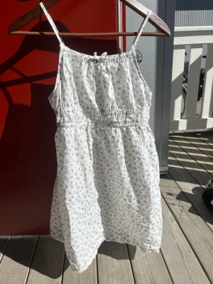Vit blommig sommarklänning med smala axelband - Söt vit kortklänning med små blå blommor och smala axelband. Klänningen har resår i midjan och är tillverkad i ett lätt bomullstyg, perfekt för varma dagar. Enkel och luftig modell som ger en avslappnad känsla. Säljs pga att den var för liten vid köp. Perfekt till sommaren🐋