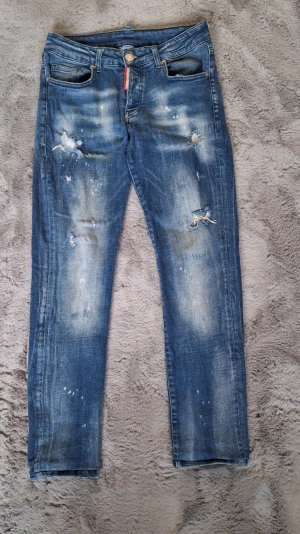 Blå jeans från Dsquared2 stl 44 - Snygga blå jeans från Dsquared2, modell 2112, med coola slitningar och blekta detaljer. Jeansen har klassisk femficksdesign, röd Dsquared2-tag vid fickan och är tillverkade i bomull med lite lycra för extra stretch. Perfekt för dig som gillar en edgy streetstil.