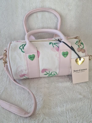 Juicy Couture rosa barrel handväska - Supergullig barrel handväska från Juicy Couture i ljusrosa och vitt med gröna detaljer och körsbärsmönster. Väskan har både handtag och axelrem, guldfärgade metalldetaljer och hjärtformade Juicy-loggor. Perfekt för dig som gillar söt och trendig stil.