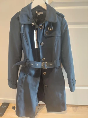 Mörkblå trenchcoat från Morgan, ny med prislapp, nypris 1200 kr - Stilren mörkblå trenchcoat från Morgan med klassisk krage, bälte i midjan och snygga spänndetaljer vid axlar och ärmslut. Knappar framtill och markerad midja ger en tidlös siluett. Perfekt för dig som vill ha en trendig och elegant look.