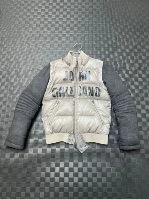 John Galliano pufferjacka grå/silver - Snygg pufferjacka från John Galliano. Jackan har hög krage, två dragkedjefickor och stor logga framtill. Perfekt för kalla dagar när du vill sticka ut med en unik 2000’s look.