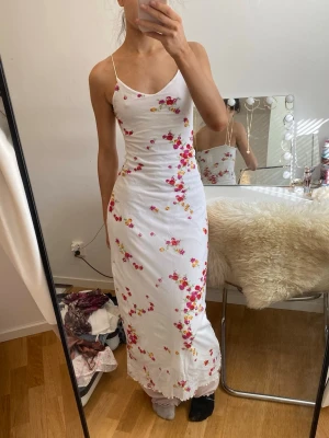 Vit blommig långklänning Chic Folie - Supersöt långklänning från Chic Folie i vitt med tunna axelband och blommigt mönster i rosa, rött och gult. Klänningen har en smickrande figurnära passform och är perfekt för sommarens alla tillfällen. Materialet känns lätt och luftigt.