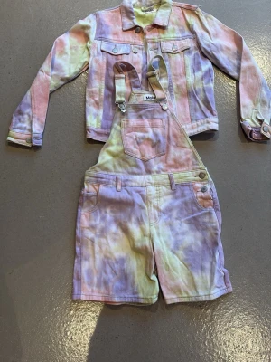 Pastellfärgade shorts med hängslen Molo - BARN: Shorts med hängslen från Molo i ett snyggt pastellfärgat batikmönster. Färgerna går i rosa, gul och lila. Modellen har klassiska fickor och knappdetaljer. Perfekt för dig som gillar färg och en lekfull stil.