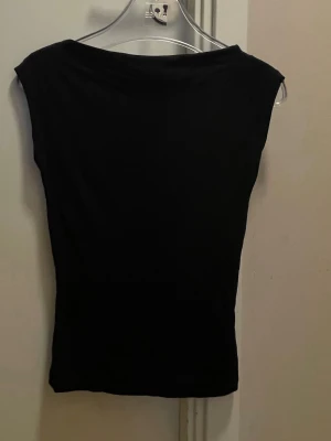 Svart ärmlös topp från Nelly - En enkel och stilren svart topp från Nelly i storlek XS. Toppen är ärmlös med en rak, något bredare halsringning och har en tight passform. Perfekt att matcha med jeans eller kjol för en clean look.